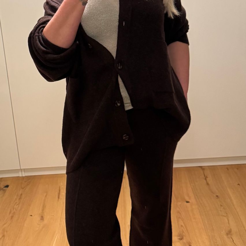 Eine Frau mit blonden Haaren, die einen schwarzen Pullover und Hosen trägt, steht auf einem Holzboden. Sie hat ein Tattoo am Arm.