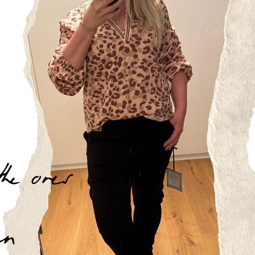 Eine Frau in einem Leopardenmuster-Blusen und schwarzen Hosen macht ein Selfie in einem Raum. Sie hält ein Handy in der rechten Hand.