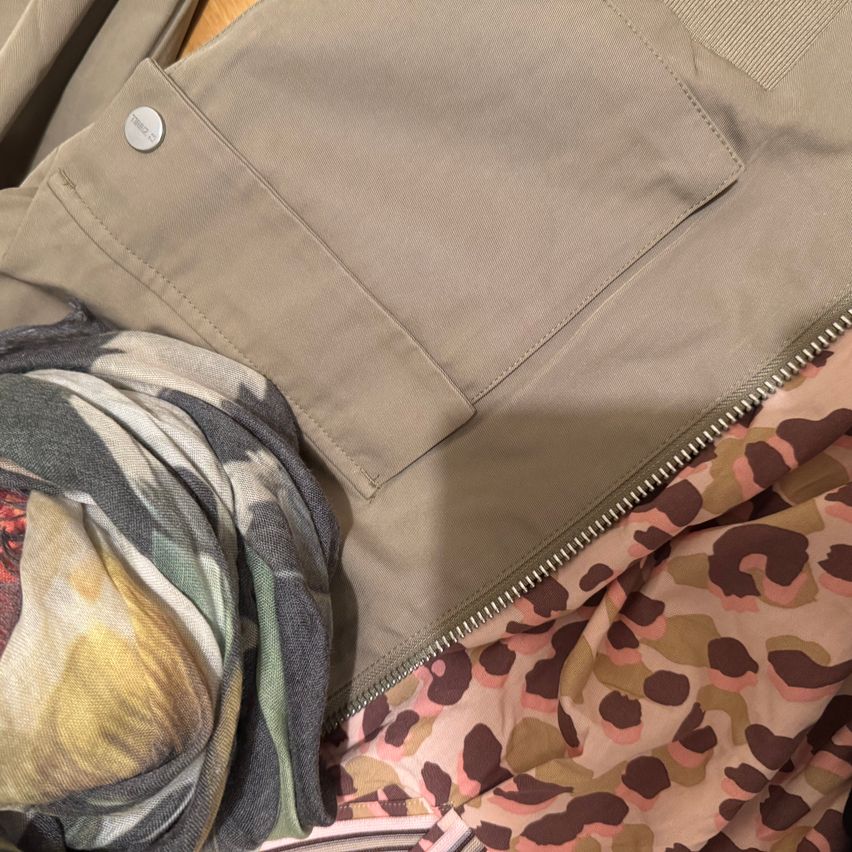 Ein beigefarbener Mantel mit einer Tarnmuster-Fütterung, einem Reißverschluss und einem Schal mit Blumenmuster ist abgebildet.