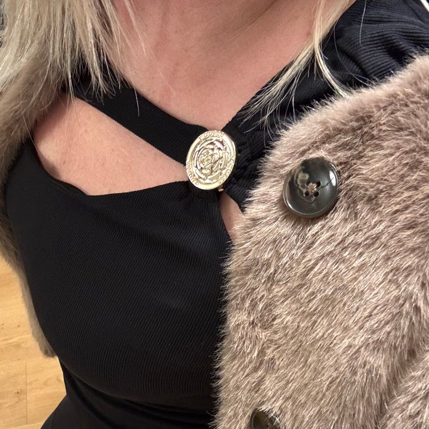 Eine Frau mit blonden Haaren trägt ein schwarzes Oberteil und einen Pelzmantel mit einem goldenen Knopf. Der Mantel hat einen schwarzen Knopf auf der rechten Seite.