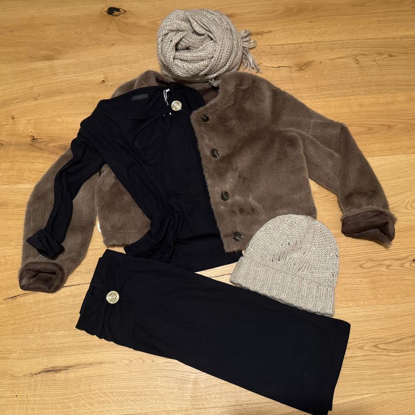 Ein schwarzes Oberteil, ein brauner Pelzmantel, eine graue Mütze und schwarze Hose auf einem Holzboden ausgelegt.