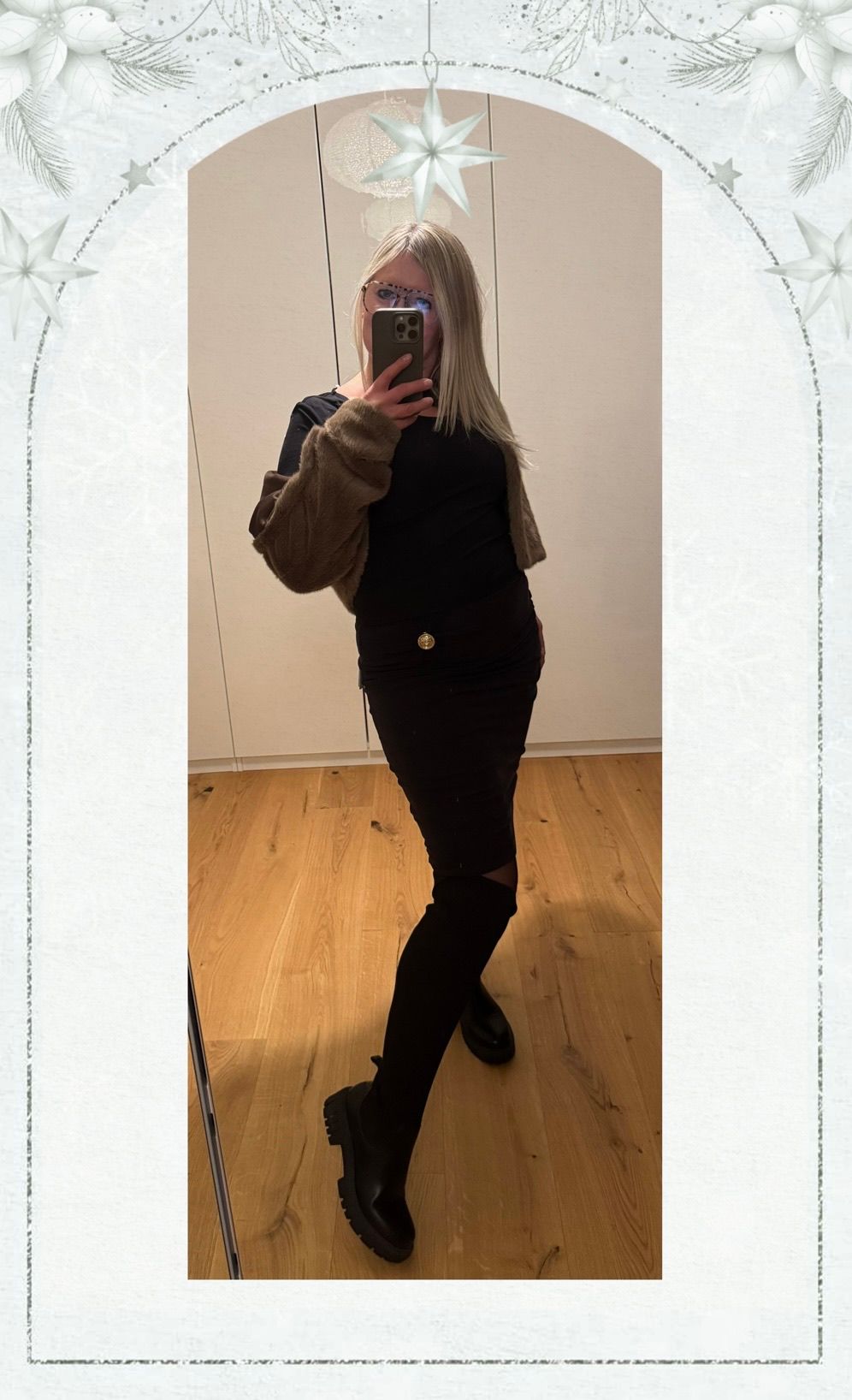 Eine Frau macht ein Selfie in Innenräumen mit einem hellen Holzboden, trägt ein schwarzes Oberteil, Hose und eine braune Jacke. Sie hält ein Smartphone.