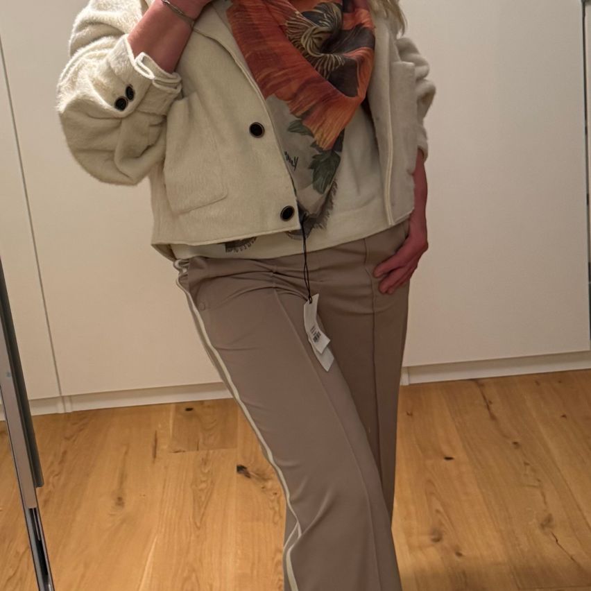 Eine Frau posiert in einer beigefarbenen Jacke und beigefarbenen Hosen, trägt einen Schal und ein Armband, mit einem weißen Etikett an der Hose, vor einer weißen Wand.
