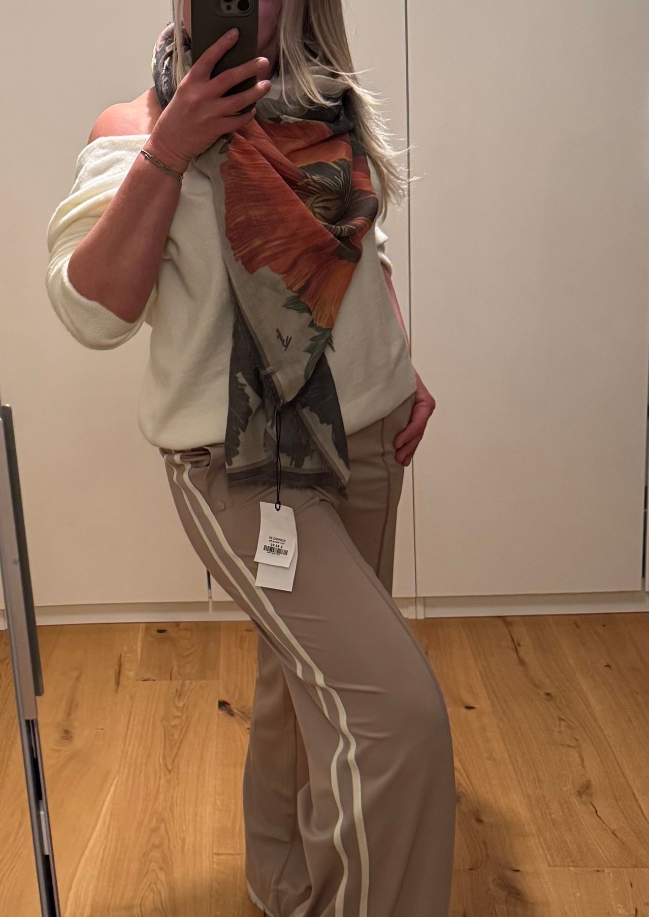 Eine Frau trägt ein beigefarbenes Oberteil, einen gemusterten Schal und beige Hosen mit weißen Streifen. Sie steht vor einem weißen Schrank und einem Holzboden.