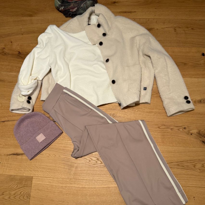 Eine weiße Bluse, eine beige Jacke, graue Hose mit weißen Streifen und eine lila Mütze liegen auf einem Holzboden.
