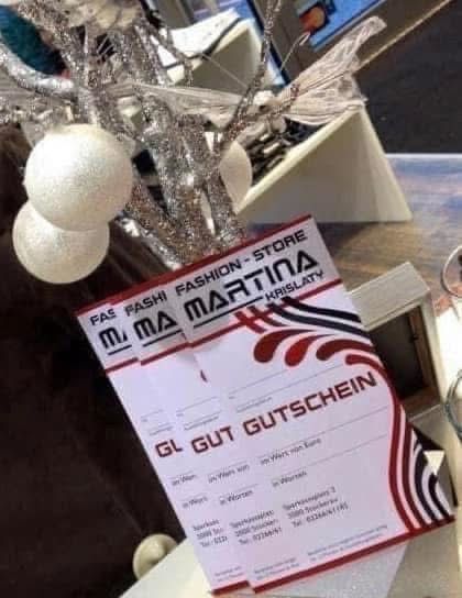 Ein Geschenkgutschein für ein Modegeschäft hängt an einem silbernen Weihnachtsbaum mit weißen Kugeln. Der Gutschein ist rot und weiß mit 'Modegeschäft Martina Krislaty' darauf geschrieben.