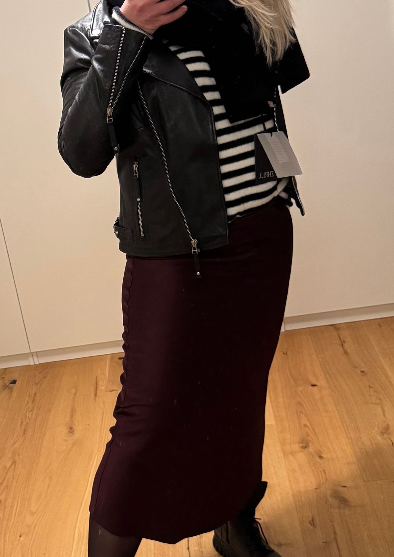 Eine Person steht in einem Raum mit Holzboden und trägt eine schwarze Lederjacke mit Reißverschlüssen und ein gestreiftes Shirt. Sie trägt auch eine purpurrote Rock.