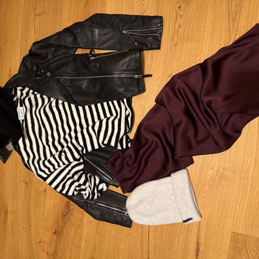 Eine schwarze Lederjacke, ein gestreiftes Hemd, eine Weinrot-Hose und eine graue Mütze auf einem Holzboden.