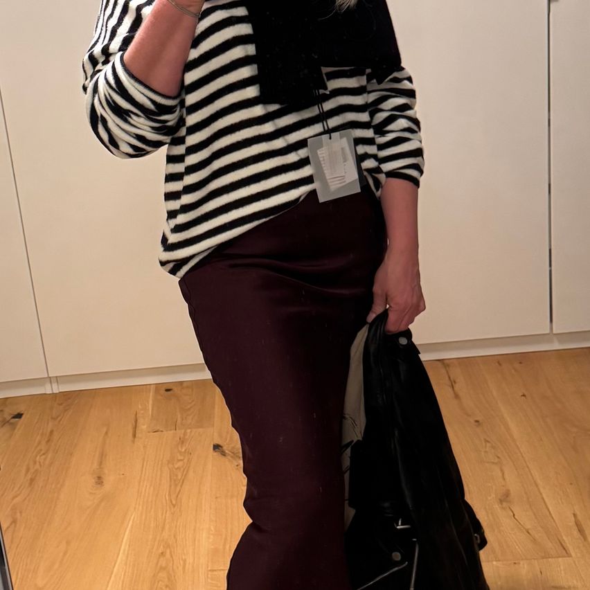 Eine Person steht drinnen und trägt ein schwarz-weiß gestreiftes Sweatshirt, dunkle Hosen und einen schwarzen Schal. Sie hält eine schwarze Jacke in der rechten Hand und steht auf einem Holzboden.