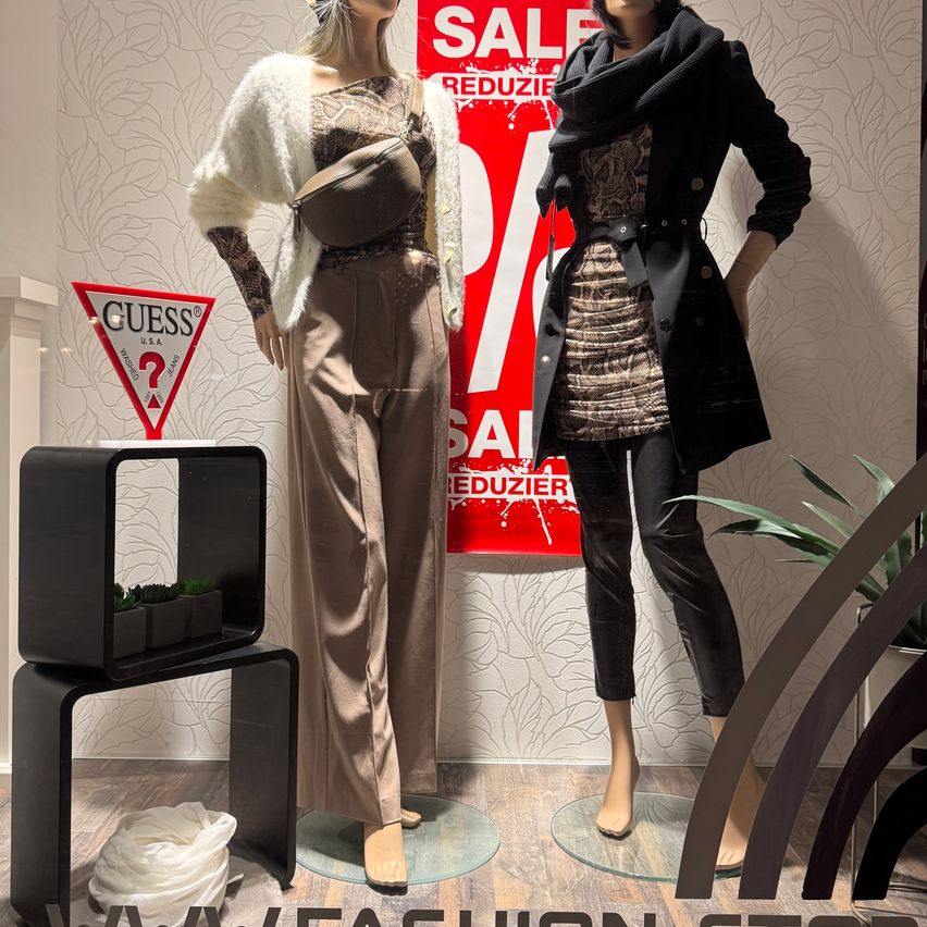 Zwei Mannequins in Guess-Kleidung stehen vor einem Verkaufsschild. Das Mannequin auf der linken Seite trägt ein beiges Outfit und eine weiße Pelzweste, während das rechte ein schwarzes Kleid und Hosen trägt. Ein dreieckiges Schild mit dem Text Guess und einem Fragezeichen steht auf einem Tisch.