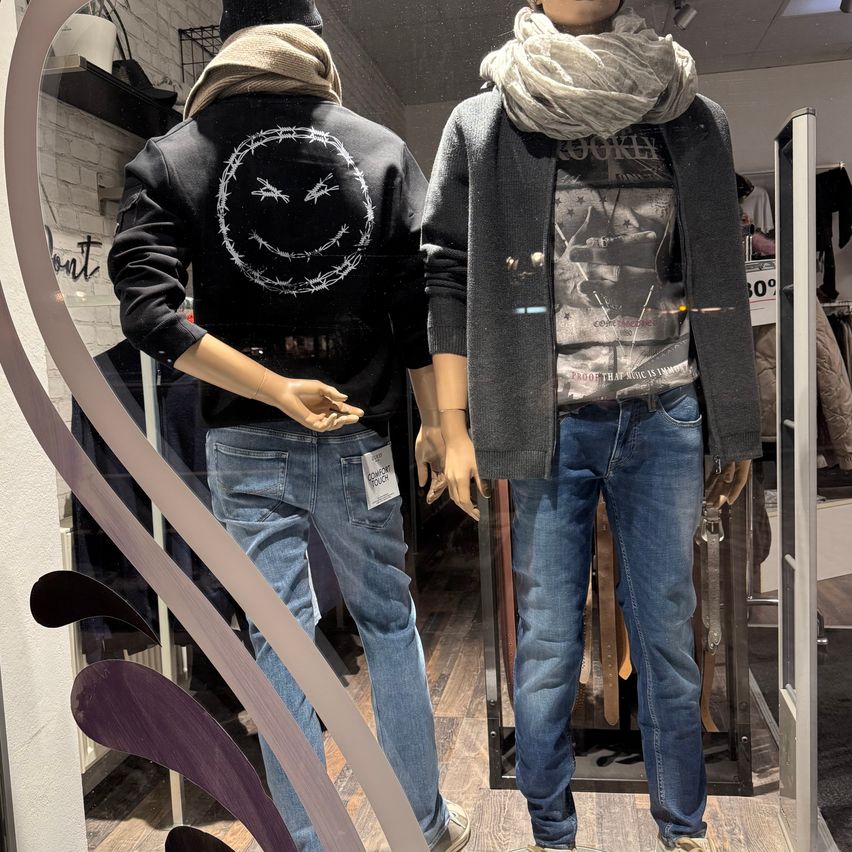 Zwei Mannequins, gekleidet in Jeans und Jacken, sind in einem Schaufenster ausgestellt. Das Mannequin auf der linken Seite trägt eine schwarze Jacke mit einem Stacheldraht-Smiley-Gesicht auf der Rückseite. Das Mannequin auf der rechten Seite trägt eine graue Jacke mit einem bedruckten T-Shirt.