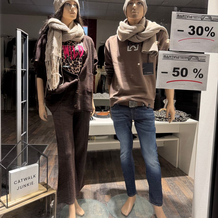 Zwei Mannequins in einem Bekleidungsgeschäft, eines trägt ein Leopardenmuster-Oberteil und braune Hose, das andere ein braunes Hemd und Jeans, beide mit Schals. Es gibt zwei Schilder mit Rabatten von 30% und 50%.