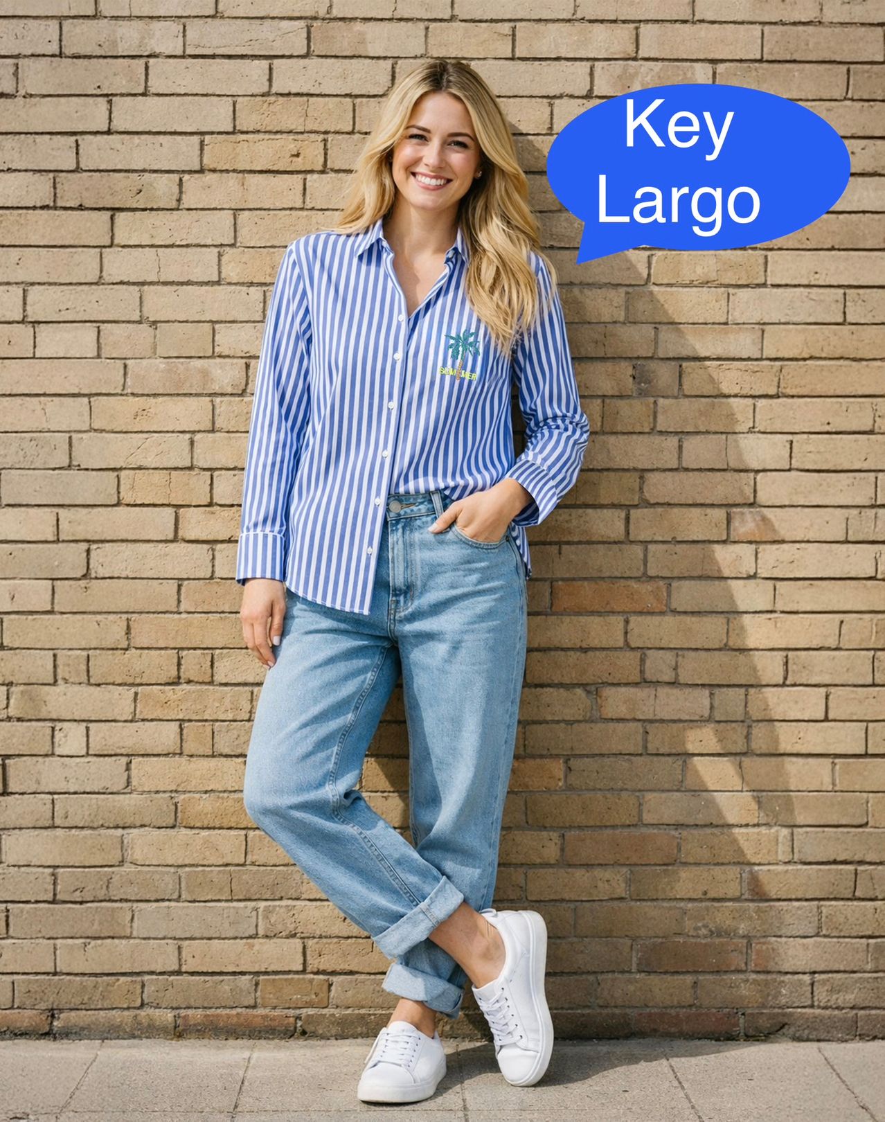 Eine Frau steht vor einer Ziegelwand in einem blau-weiß gestreiften Hemd, blauer Jeans und weißen Sneakern. Sie hat eine Hand in der Tasche.