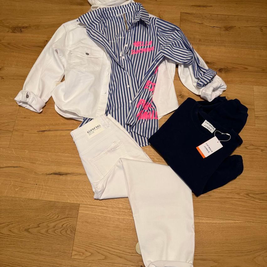 Ein weißes Hemd, weiße Jeans und ein marineblauer Sweatshirt mit weißem Etikett liegen auf einem Holzboden.