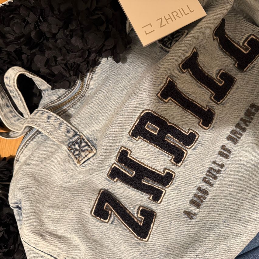 Eine Nahaufnahme eines grauen Sweatshirts mit dem Wort 'ZHR1LL' in fetten, schwarzen und goldenen Buchstaben auf der Vorderseite. Es hat einen Reißverschluss und ein Etikett mit der Aufschrift 'ZHR1LL'.