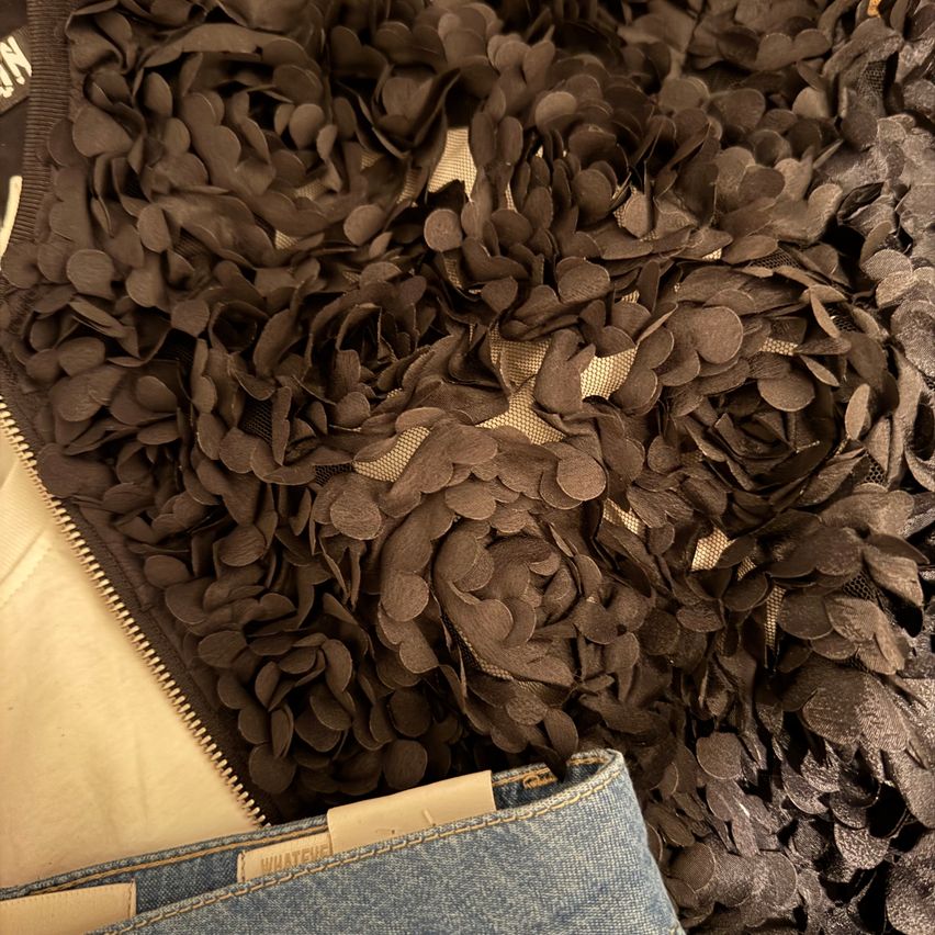 Eine Nahaufnahme eines braunen Paillettenjacken mit Reißverschluss und einem darunter sichtbaren Jeansrock. Die Jacke hat ein Blumen-Paillettenmuster.