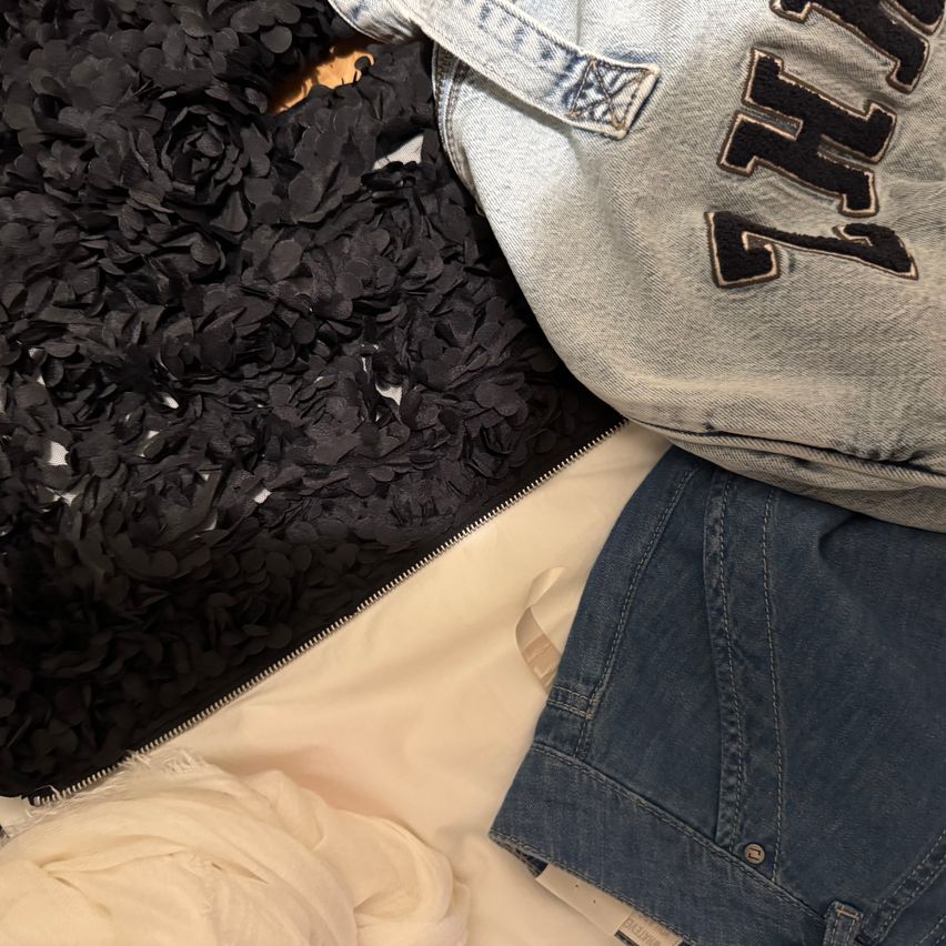 Eine graue Tasche mit dem Wort 'NY' in schwarzen Buchstaben liegt auf einem weißen Tuch. Daneben ist eine Jeansjacke mit einem Logo auf der linken Seite. Schwarzer Stoff mit einem Blumendesign ist ebenfalls auf dem weißen Tuch.