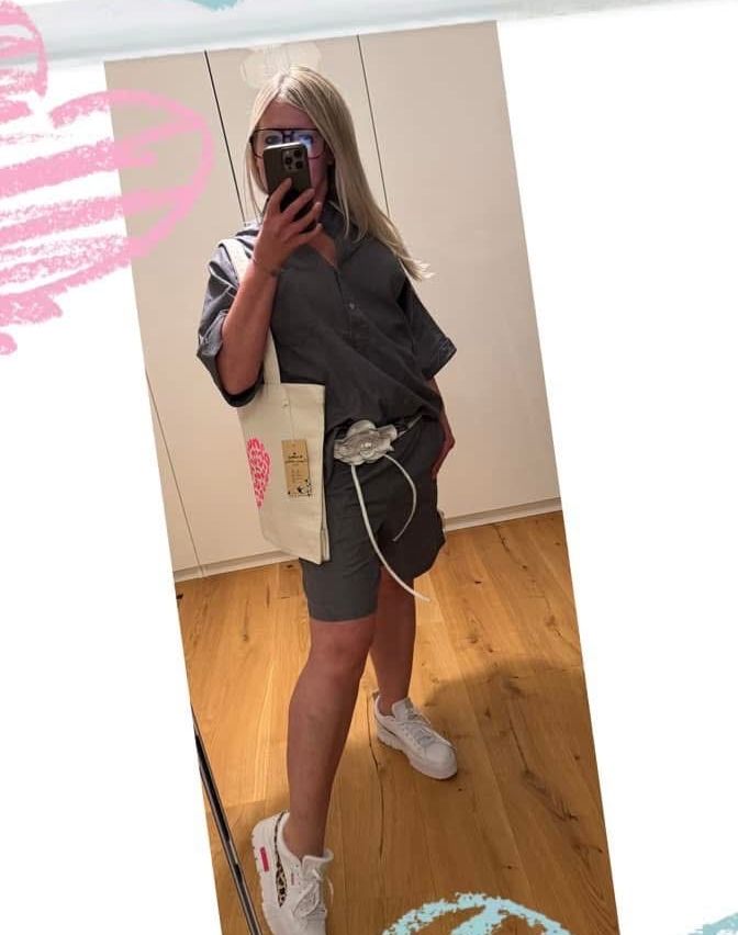 Eine Frau in einem grauen Shirt und Shorts macht ein Selfie mit ihrem Handy, während sie eine weiße Tasche mit einem roten Logo hält. Sie steht auf einem Holzboden mit einem weißen Rand.
