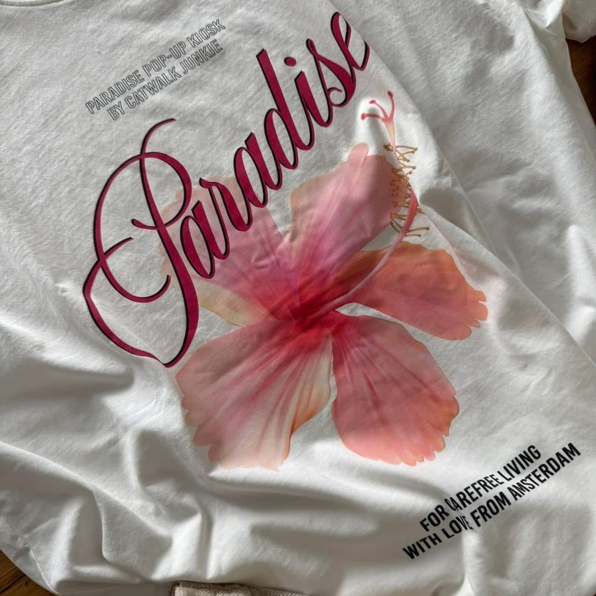 Ein weißes T-Shirt mit dem Wort 'Paradise' in pink und roten Buchstaben, mit einem rosa Blumendesign. Es trägt auch 'Paradise Pop-Up Kiosk by Catwalk Junkie' und 'Für ein Refree Leben mit Liebe aus Amsterdam.'