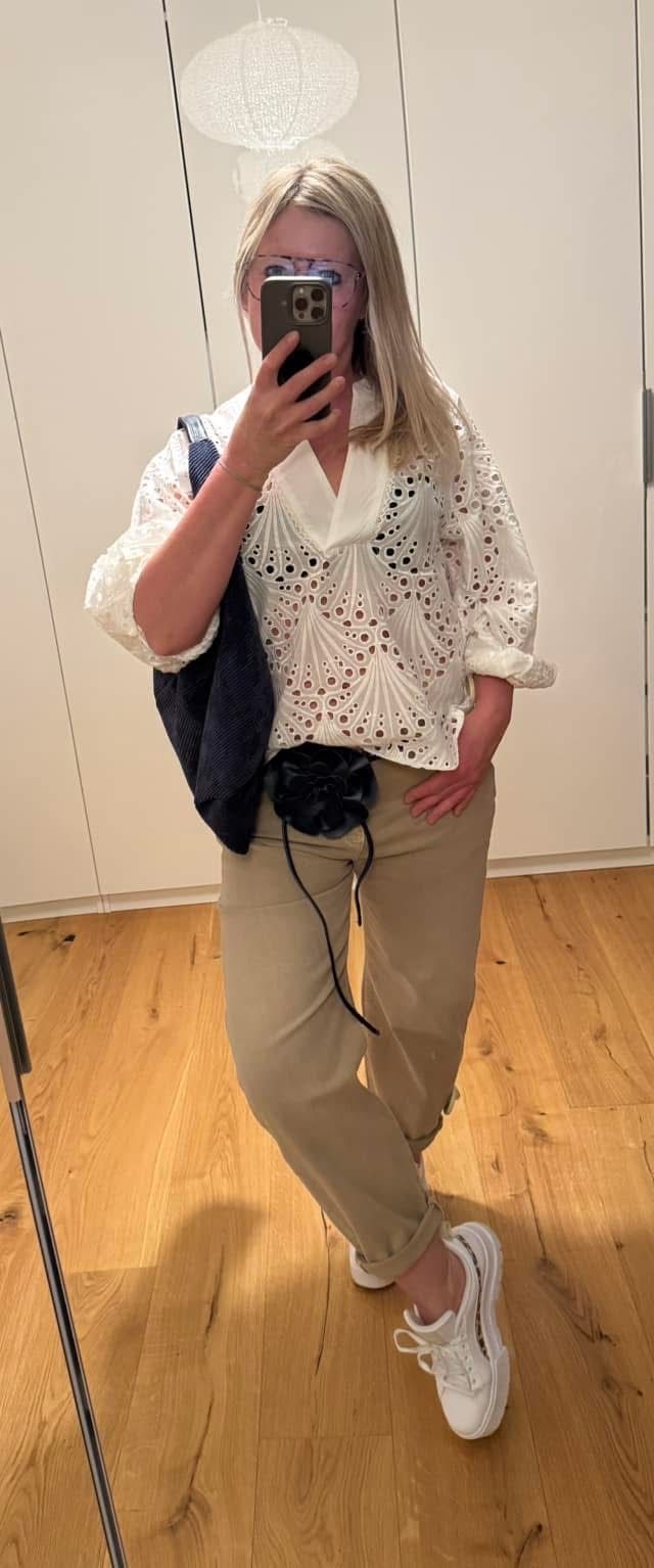 Eine Frau macht ein Selfie, trägt eine weiße Bluse mit Spitzendetails, beige Hose und eine schwarze Tasche mit einer schwarzen Blume am Gürtel.