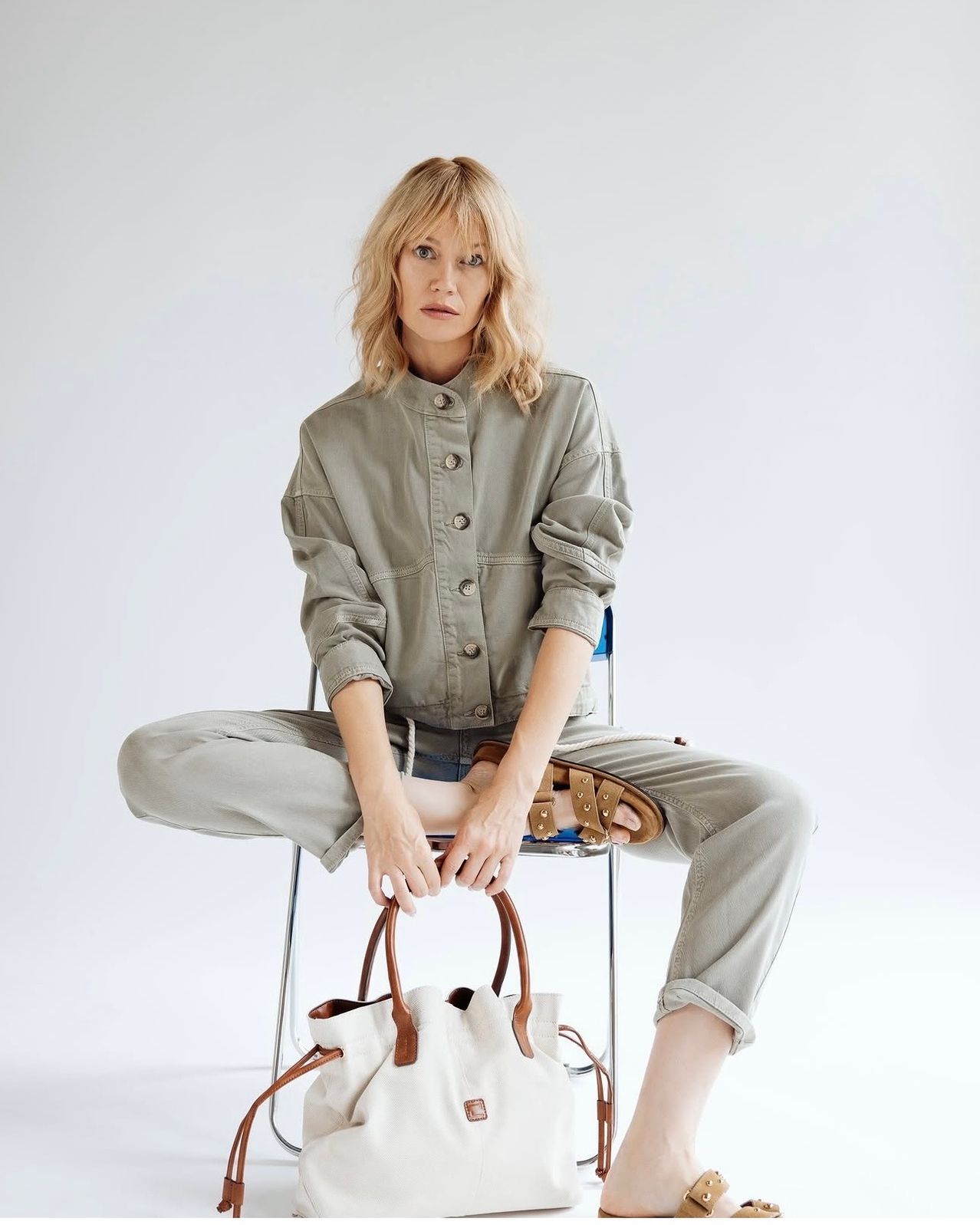 Eine Frau mit blondem Haar sitzt auf einem Stuhl und hält eine braune Handtasche und ein beiges Shirt und eine Hose.