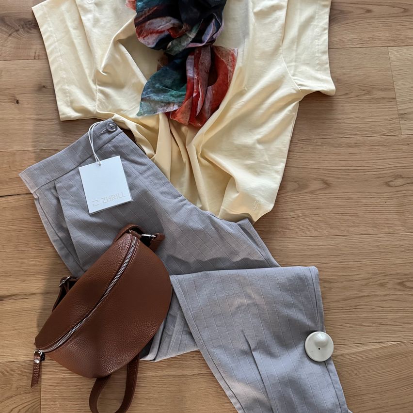 Ein beigefarbenes Hemd, ein Schal, graue Hosen, ein brauner Leder-Fanny-Pack und ein weißer Knopf liegen auf einem Holzboden.