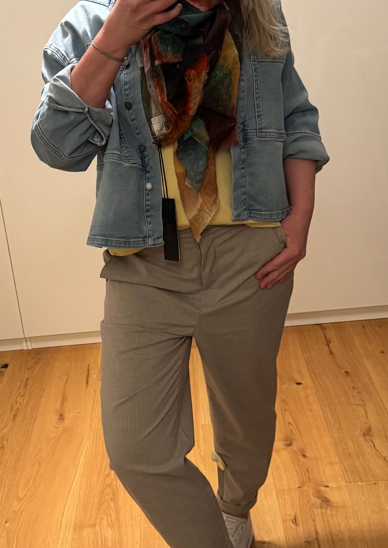 Eine Person steht mit einer Hand in der Tasche, trägt eine Jeansjacke, ein gelbes Hemd und graue Hose. Ein Schal ist um ihren Hals gelegt.