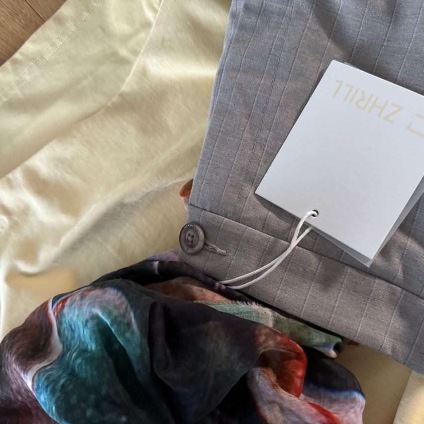 Ein grauer Anzugsakkordeon mit einem Knopf und einem Schild mit der Aufschrift 'ZHZ 1718' liegt auf einem cremefarbenen Stoff mit einem farbenfrohen Krawatte darunter.