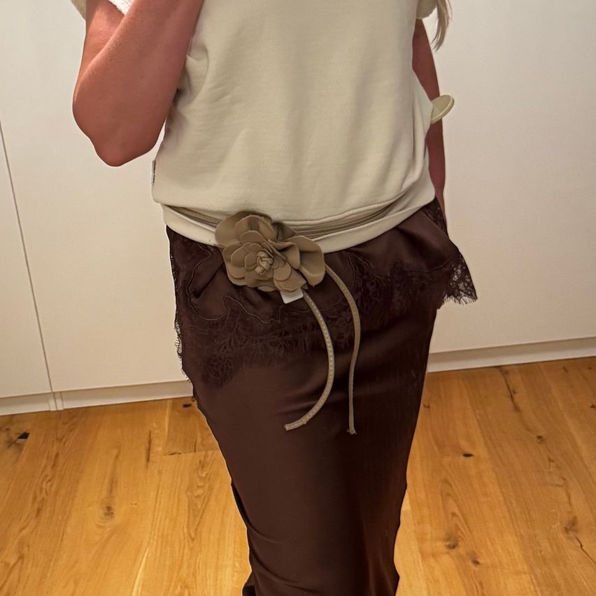 Eine Frau trägt ein beigefarbenes Oberteil und einen braunen Rock mit Blumengürtel. Sie steht auf einem Holzboden.
