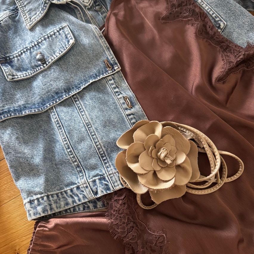 Eine Jeansjacke, ein braunes Seidenkleid und eine beige Blumenbrosche liegen auf einer Holzoberfläche.