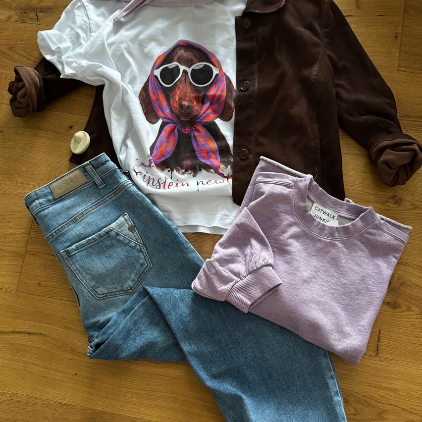 Ein weißes T-Shirt mit einem Dackel, der eine Sonnenbrille trägt, eine braune Jacke, blaue Jeans und ein lila Sweatshirt liegen auf einem Holzboden.