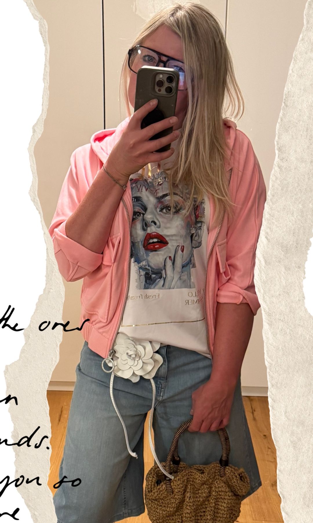A woman in a pink hoodie with a floral waist bag takes a selfie. She wears a white t-shirt with a picture of a woman's face.