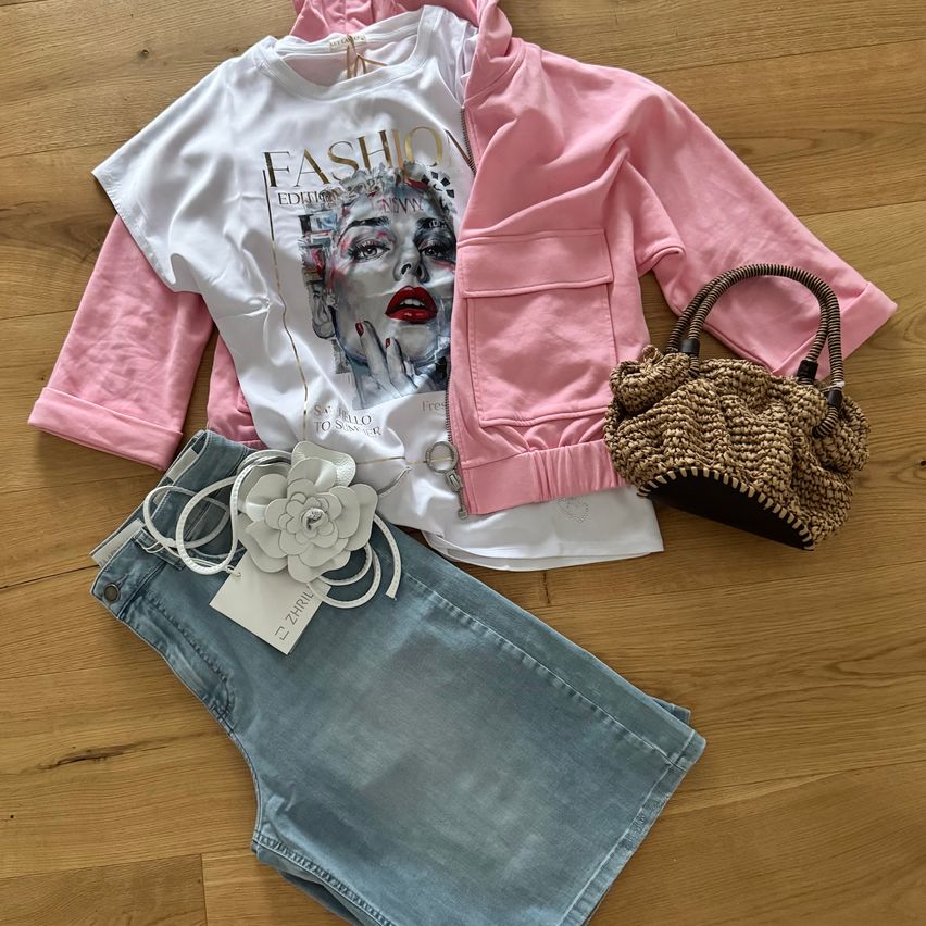 A white t-shirt with a woman's portrait, pink jacket, denim shorts, and a brown handbag are laid on a wooden floor.
