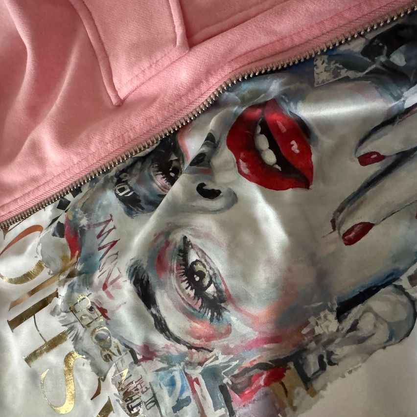 A pink hoodie with a zipper displaying an abstract painting of a woman's face, featuring bold red lips and striking eyes, with the word 'SISTER' written in gold on the bottom left.