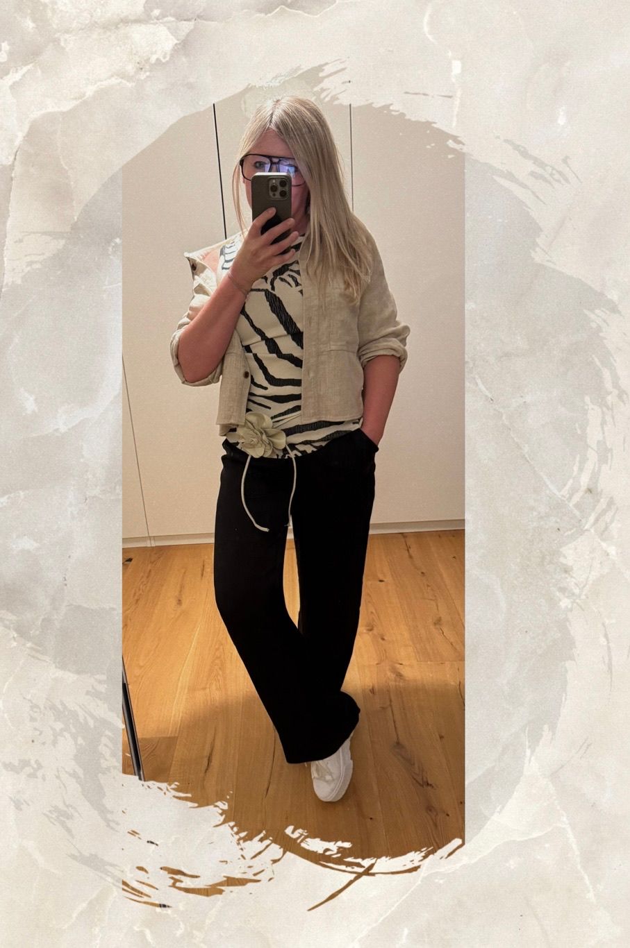 A woman is taking a selfie in front of a mirror. She wears a beige jacket over a zebra-print shirt and black pants with a white ribbon at the waist.