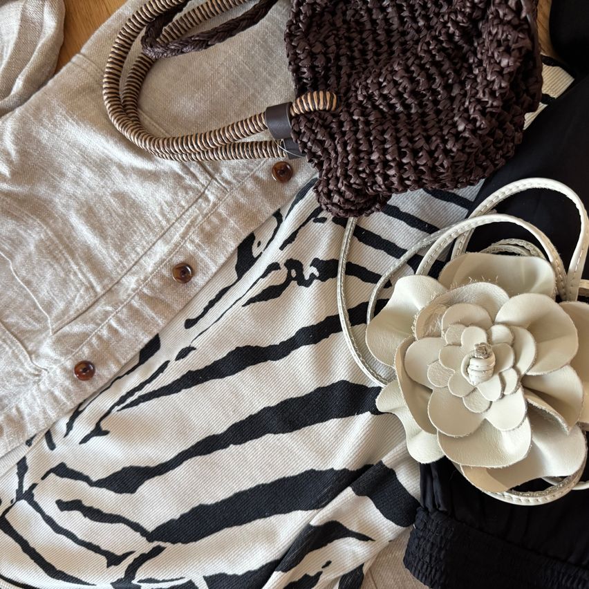 A brown woven handbag, a white flower accessory, a white striped shirt, and a white jacket are on a wooden surface.
