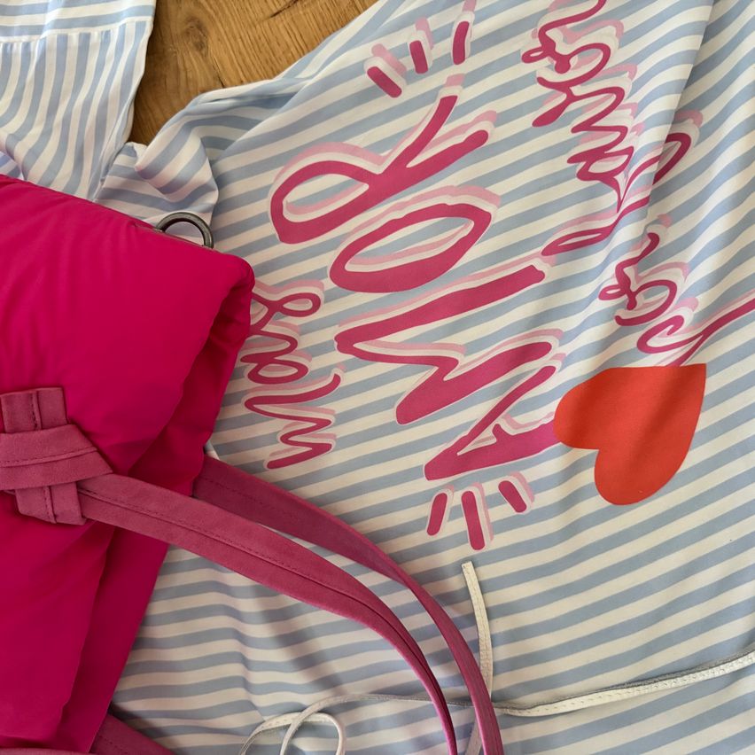 Ein rosa Rucksack liegt auf einem blau-weiß gestreiften Stoff mit dem Wort 'LIEBE' in Rosa geschrieben.