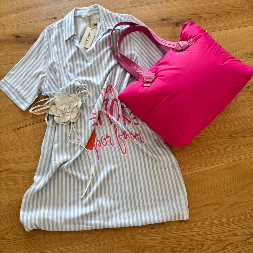 Ein blau-weiß gestreiftes Hemd mit pinker Schrift und ein rosa Kissen liegen auf einem Holzboden. Eine pinke Handtasche mit Henkel befindet sich neben dem Hemd.