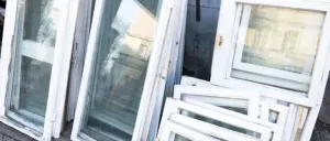 Eine Sammlung alter weißer Fensterrahmen mit Glasscheiben, die auf einem Bürgersteig gestapelt sind, einige zeigen Reflexionen von Gebäuden und Bäumen.