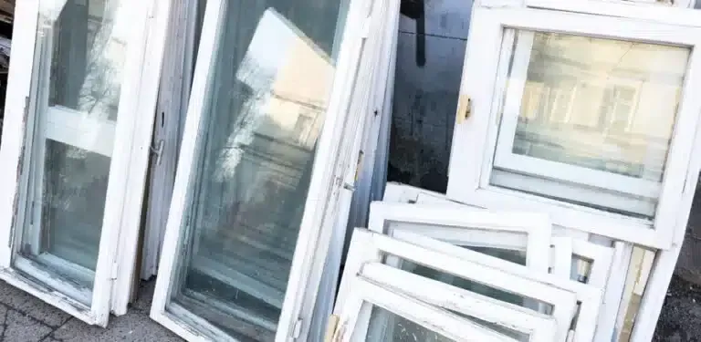 Eine Sammlung alter weißer Fensterrahmen mit Glasscheiben, die auf einem Bürgersteig gestapelt sind, einige zeigen Reflexionen von Gebäuden und Bäumen.