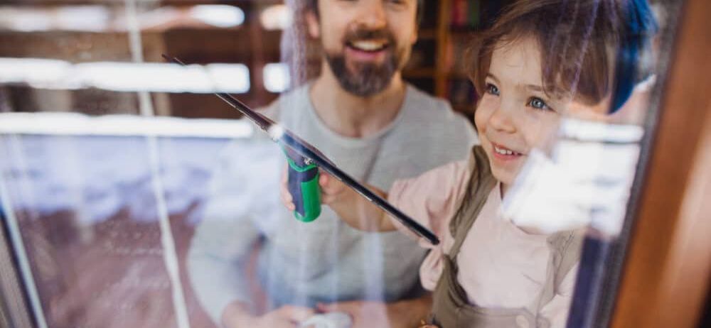 Ein Mann und ein junges Mädchen putzen ein Fenster mit einem Gummizug und einem Tuch und lächeln sich an.