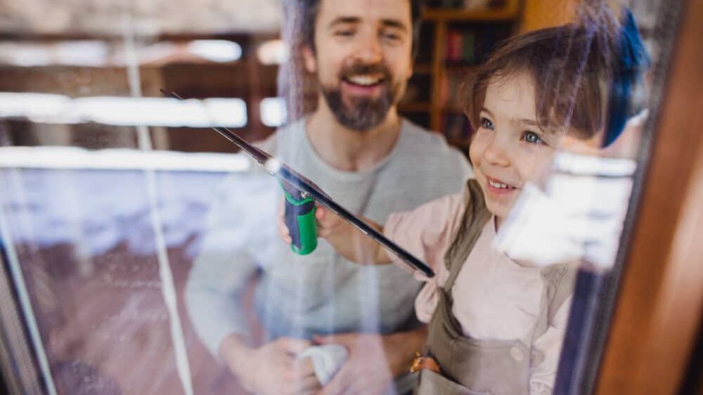 Ein Mann und ein junges Mädchen putzen ein Fenster mit einem Gummizug und einem Tuch und lächeln sich an.