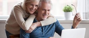 Ein älteres Paar lächelt, während es an einem Schreibtisch mit einem Laptop sitzt. Die Frau steht hinter dem Mann, legt ihre Hände auf seine Schultern und lächelt.