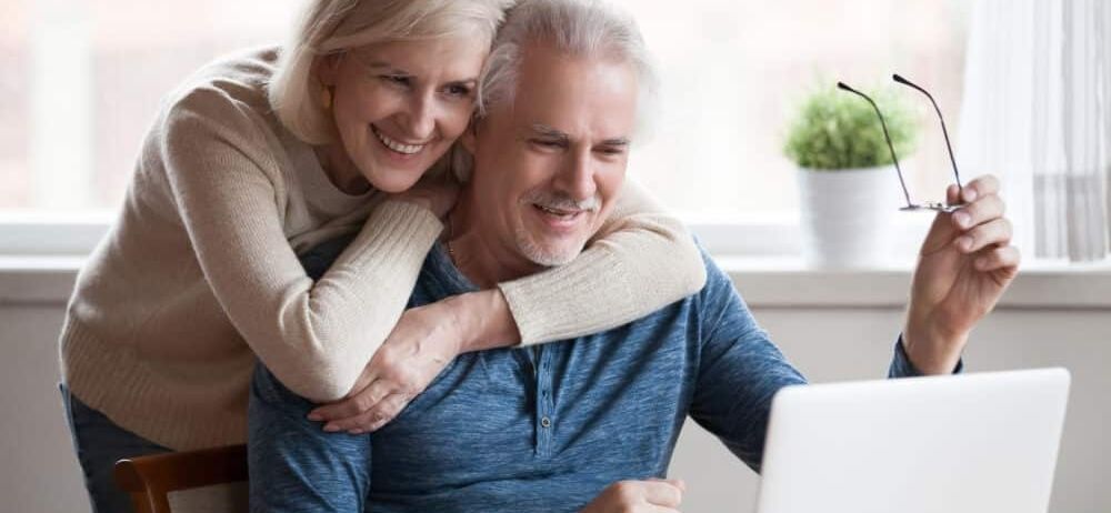 Ein älteres Paar lächelt, während es an einem Schreibtisch mit einem Laptop sitzt. Die Frau steht hinter dem Mann, legt ihre Hände auf seine Schultern und lächelt.