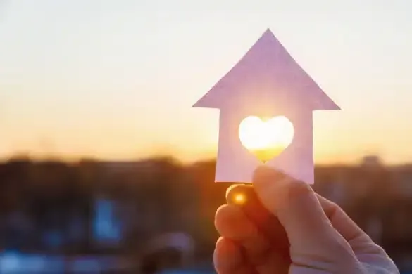 Eine Hand hält ein herzförmiges Ausschnitt in einer papiernen Haussilhouette mit Sonnenlicht, das bei Sonnenuntergang leuchtet.