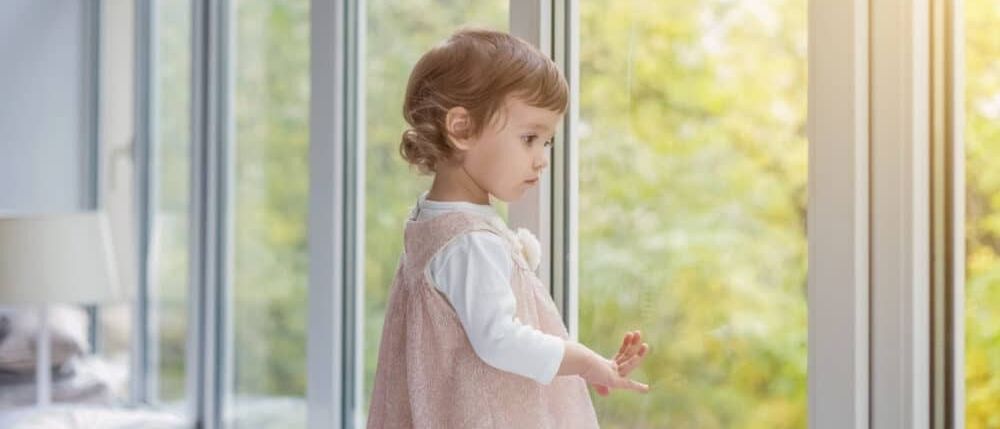 Ein kleines Mädchen in einem rosa Kleid steht vor einem Fenster und blickt neugierig nach draußen. Das Fenster reflektiert die Bäume und das Sonnenlicht und schafft eine ruhige Atmosphäre.
