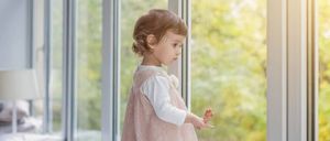 Ein kleines Mädchen in einem rosa Kleid steht vor einem Fenster und blickt neugierig nach draußen. Das Fenster reflektiert die Bäume und das Sonnenlicht und schafft eine ruhige Atmosphäre.