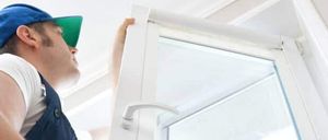 Ein Mann in blauer Arbeitskleidung und einer blauen Kappe öffnet ein Fenster an einer weißen Wand.