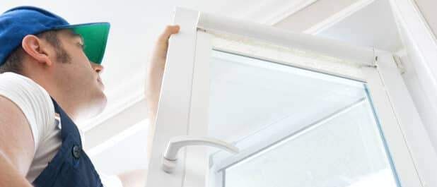 Ein Mann in blauer Arbeitskleidung und einer blauen Kappe öffnet ein Fenster an einer weißen Wand.