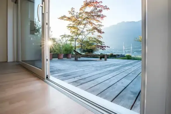 Ein Blick durch eine offene Tür auf eine Holzterrasse mit einer Liege und Bäumen.